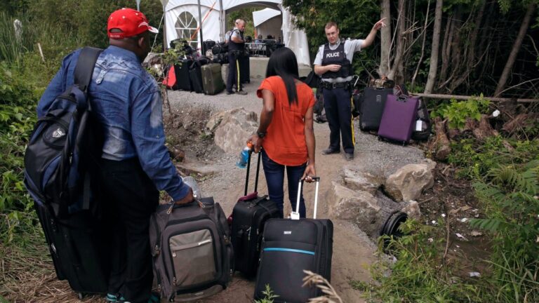 In this Aug. 7, 2017 file photo, a Royal Canadian Mounted Police officer informs a migrant couple of the location of a legal border station, shortly before they illegally crossed from Champlain, N.Y., to Saint-Bernard-de-Lacolle, Quebec, using Roxham Road. (AP Photo/Charles Krupa)