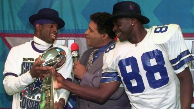 Dallas Cowboys cornerback Deion Sanders, left, and running back Michael Irvin (88) share the Vince Lombardi trophy as NBC commentator Greg Gumbel interviews the two after Super Bowl XXX in Tempe, Ariz., Sunday, Jan. 28, 1996. (AP Photo/Ron Heflin, File)